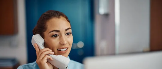Nurse answering a phone in front of a computer at a desk in a medical office.