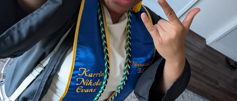A person wearing undergraduate graduation cap and gown, with some cords. The person is making a rock-n-roll hand sign