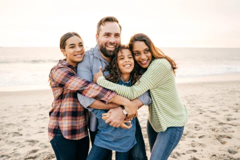 A family of four smiling and hugging on a beach