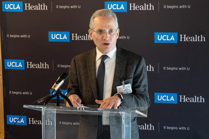 White man with short grey hair and glasses in a suit speaking in front of a clear podium with "UCLA Health, It begins with U" background.