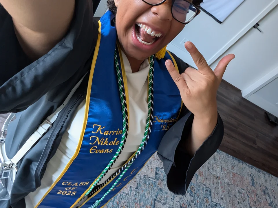 A person wearing undergraduate graduation cap and gown, with some cords. The person is making a rock-n-roll hand sign