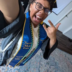 A person wearing undergraduate graduation cap and gown, with some cords. The person is making a rock-n-roll hand sign