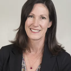 A white woman with dark brown mostly straight hair to shoulders. She has brown eyes and is smiling. She is wearing a black business jacket, the blouse is mostly cut off but can see some blue and brown. She is wearing a red pendant. 