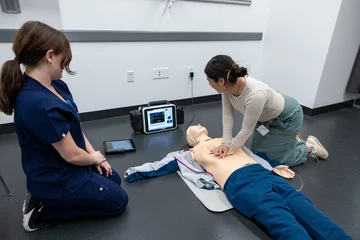 Two students are practicing CPR Compressions on a simulation adult dummy. One is watching the other practice compressions and observing the monitor.