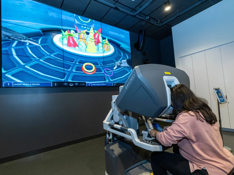 A woman is sitting down with her face in a machine. Her hands are using the machine joysticks that match as surgery tools on the screen in front of her. She is performing a simulated surgery game via this machine.